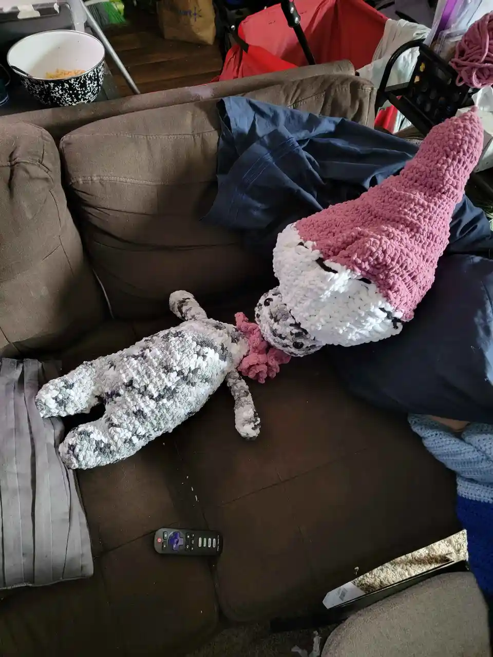 A crocheted fox plushie wearing a pink hat is lying on a brown couch, appearing to bite or chew on a smaller, gray-and-white…