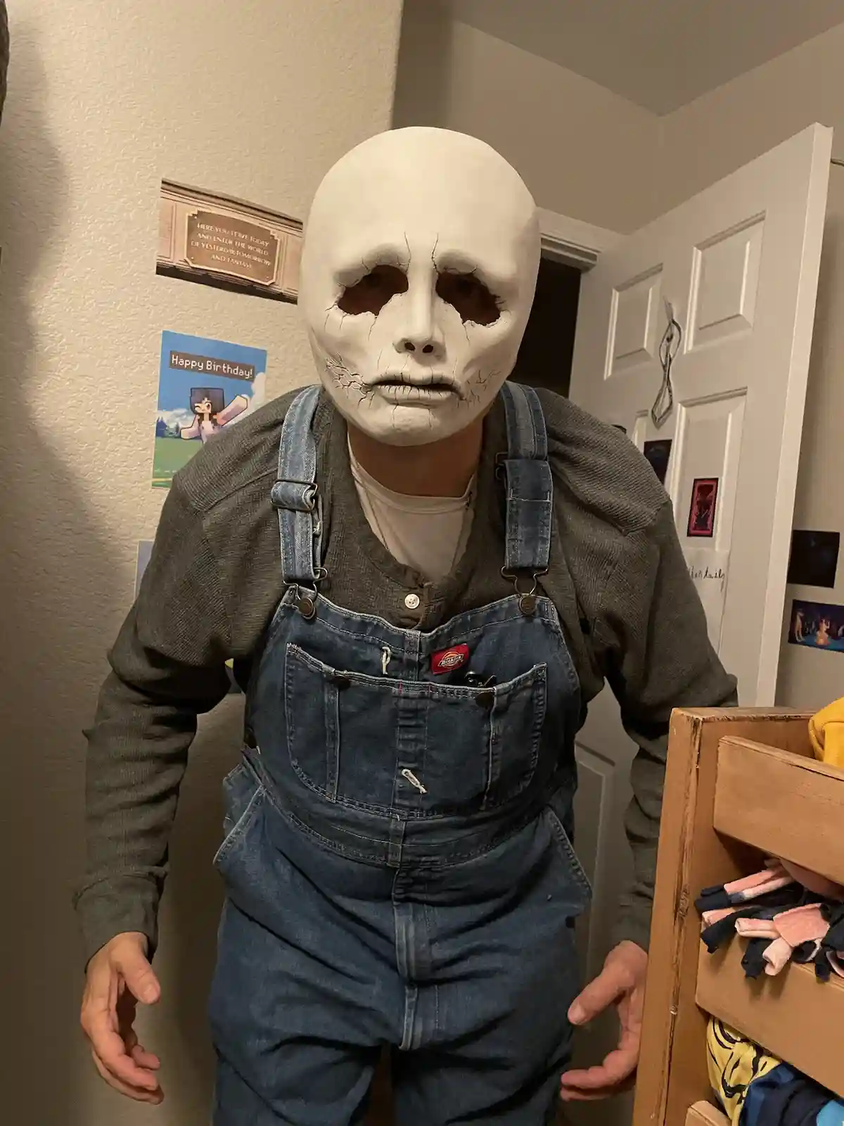 A person wearing a white, cracked, skull-like mask and blue denim overalls stands indoors, with a 'Happy Birthday!' poster…