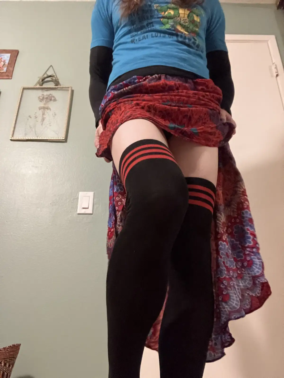 A person is shown from the waist down, lifting a red and blue patterned skirt to reveal black knee-high socks with red…