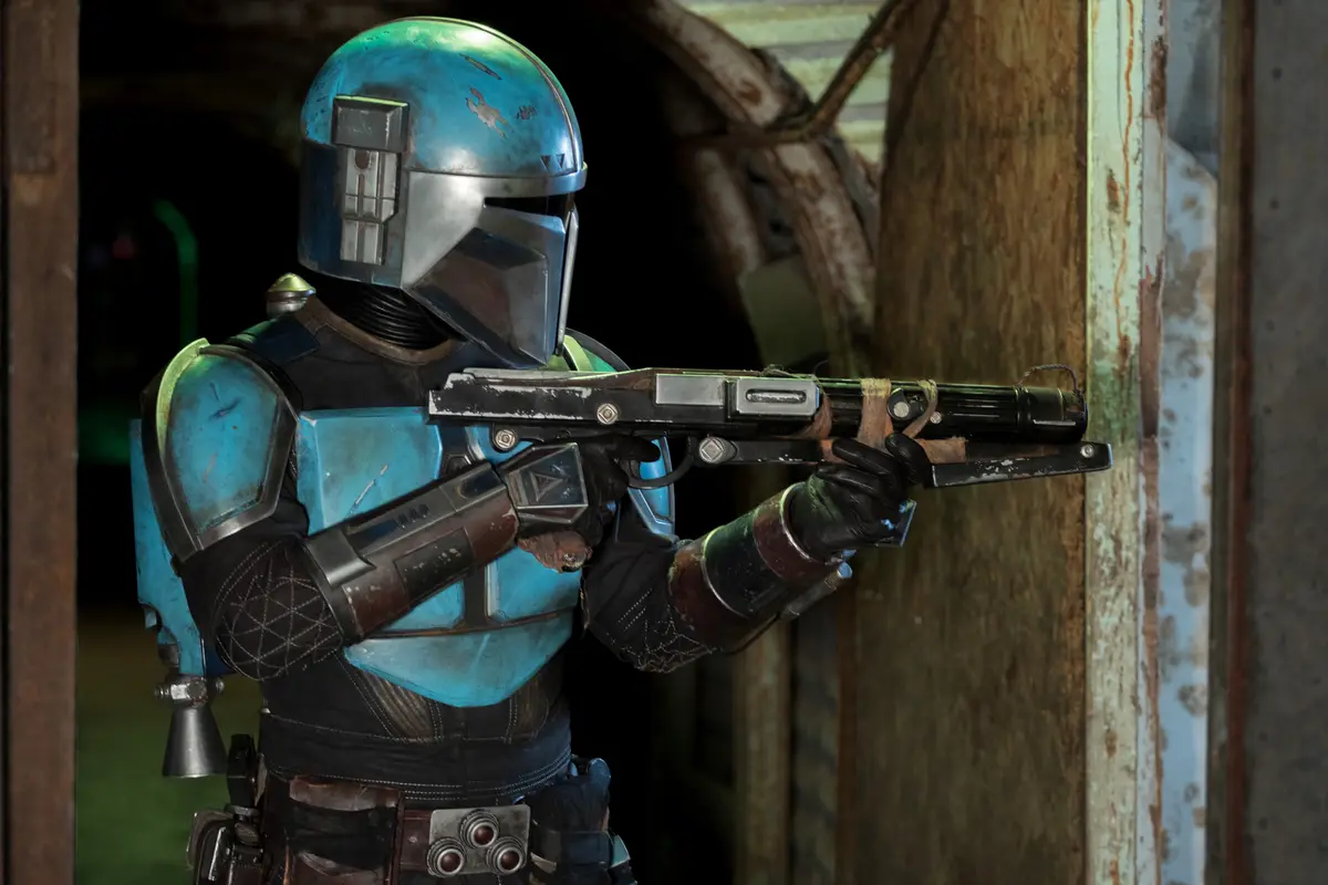 A character in blue and black Mandalorian armor, holding a blaster rifle, stands in a doorway with a weathered, rusty metal…