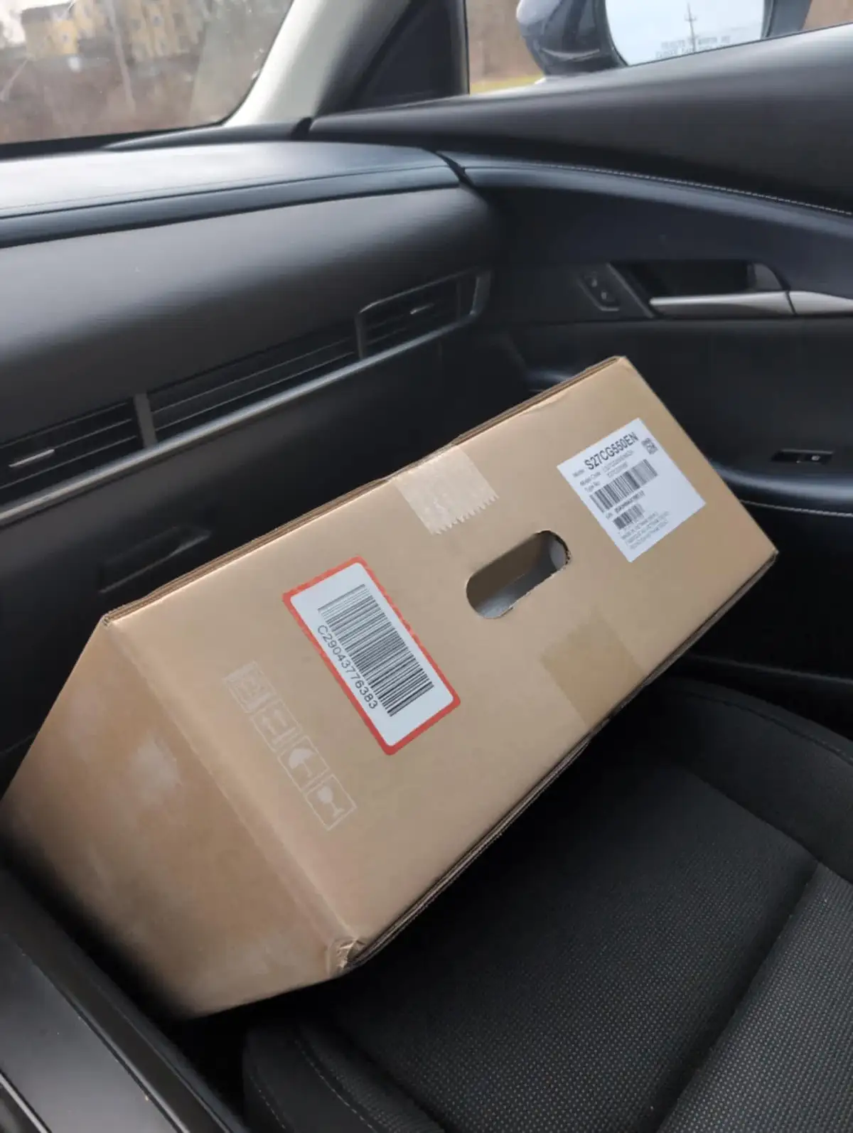A plain brown cardboard shipping box with barcodes and handling symbols sits awkwardly on the passenger seat of a modern…