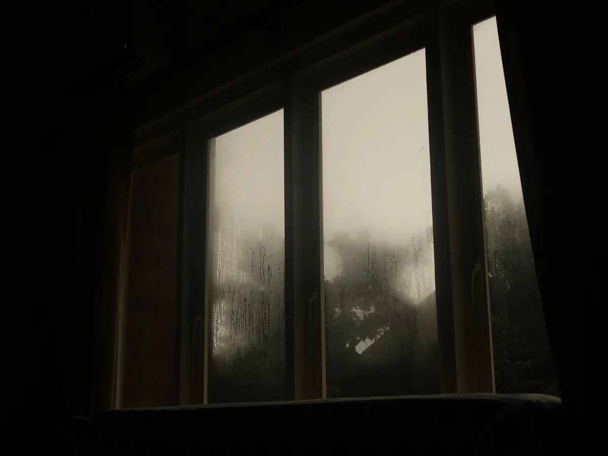 A dimly lit indoor shot looking out a multi-pane window covered in rain streaks and condensation, with a blurry outdoor…