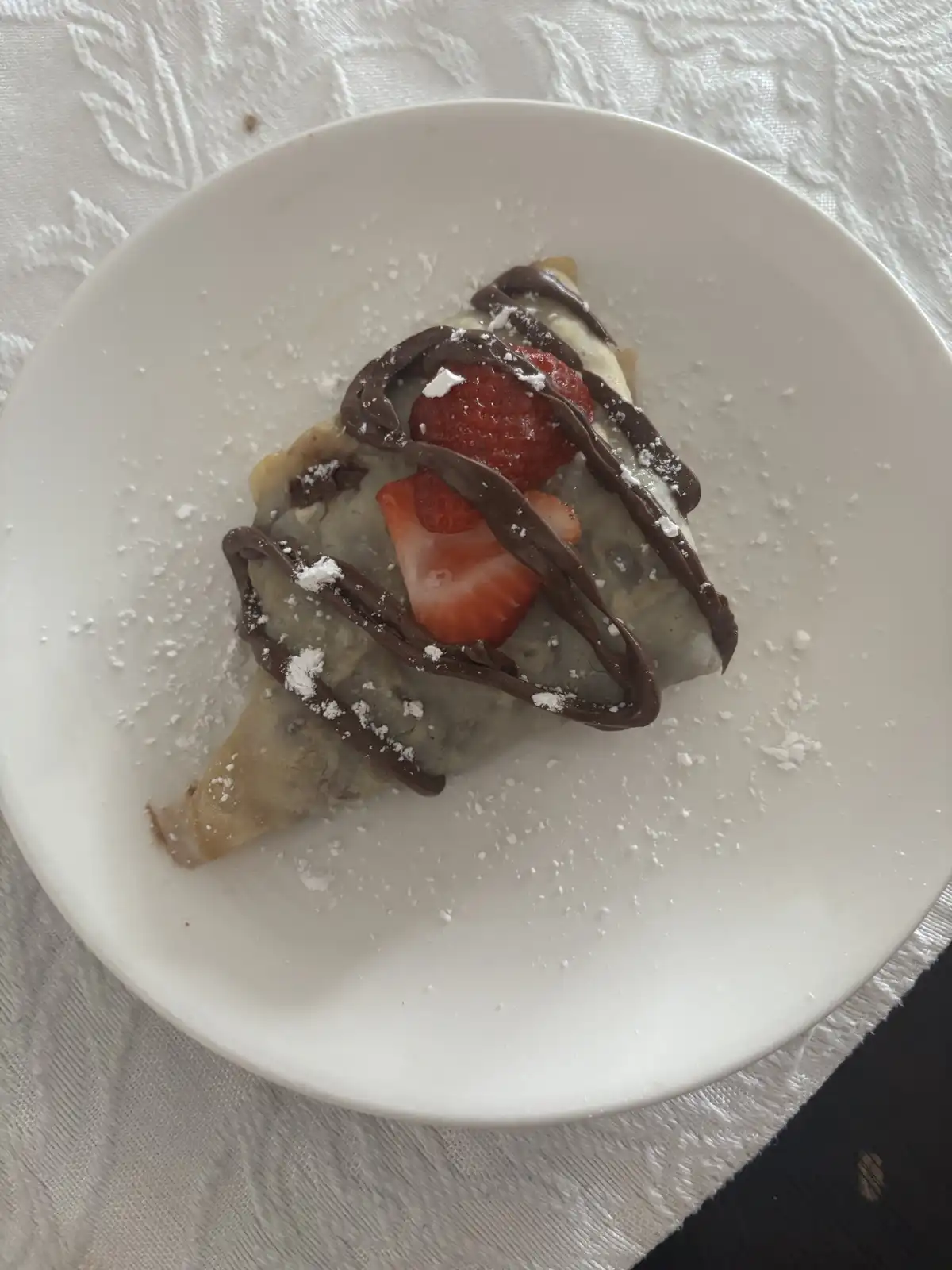 A folded crepe on a white plate, drizzled with chocolate sauce, topped with sliced strawberries and dusted with powdered…