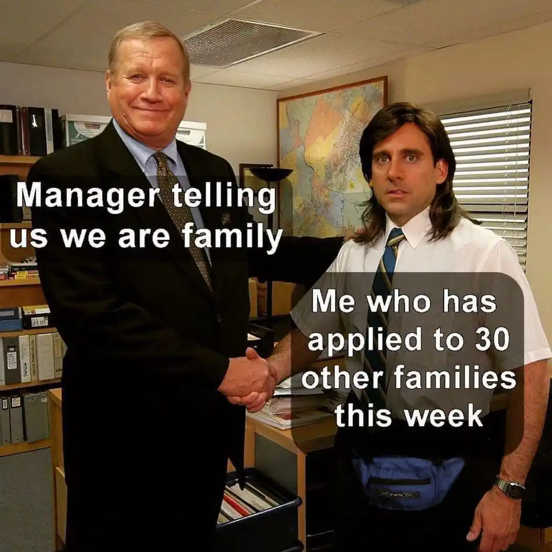 Two men in an office setting, one in a suit smiling while shaking hands with the other, who has long hair, a blue fanny…