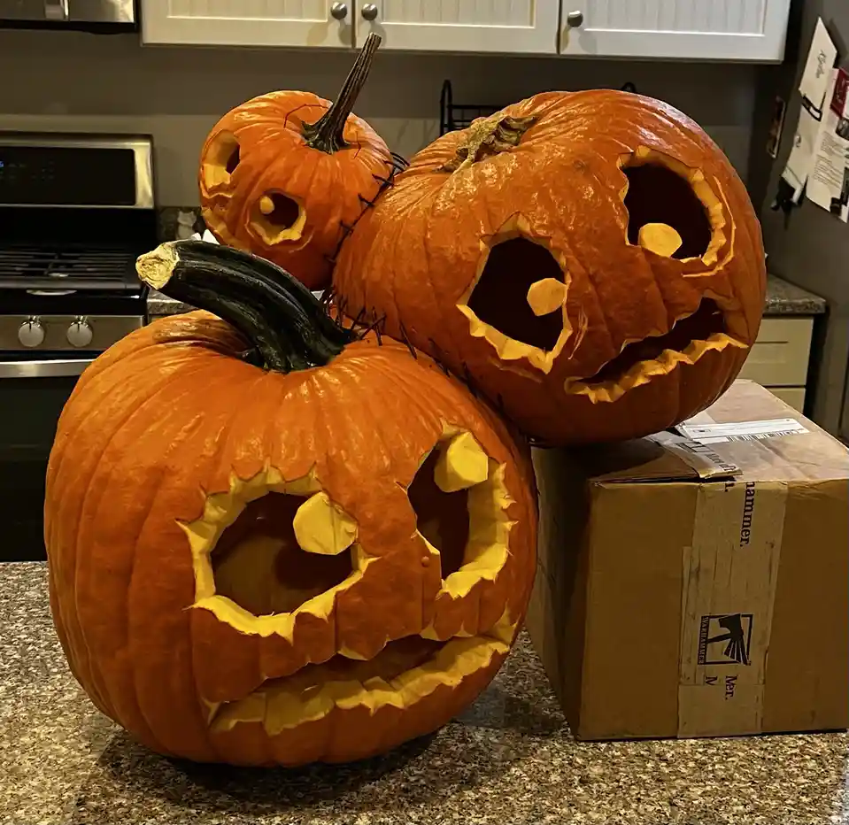 Three carved jack-o'-lanterns sit on a kitchen counter, with the largest one in front and two smaller ones behind it. The…