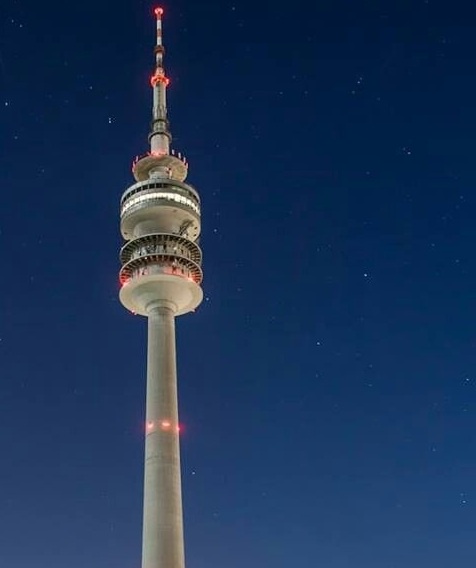 A tall, white observation tower with red lights at its top and along its shaft stands against a dark, starry night sky. The…