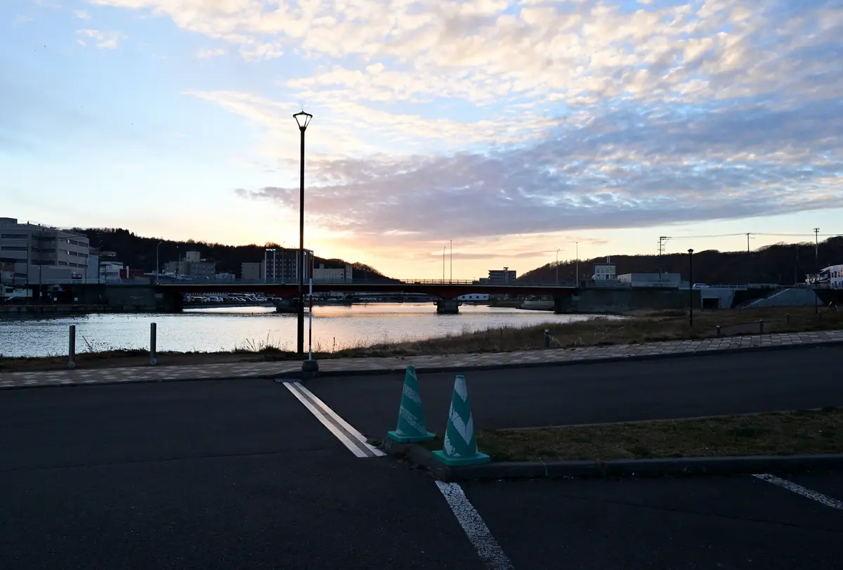 A serene, slightly dim urban riverside scene at dawn or dusk, featuring a paved road with two weathered green-and-white…