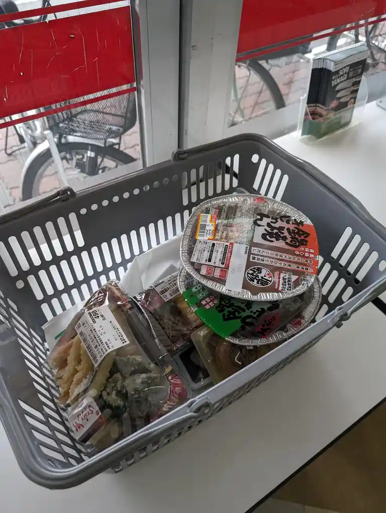 A gray plastic shopping basket sits on a white counter, filled with Japanese convenience store food items including a…