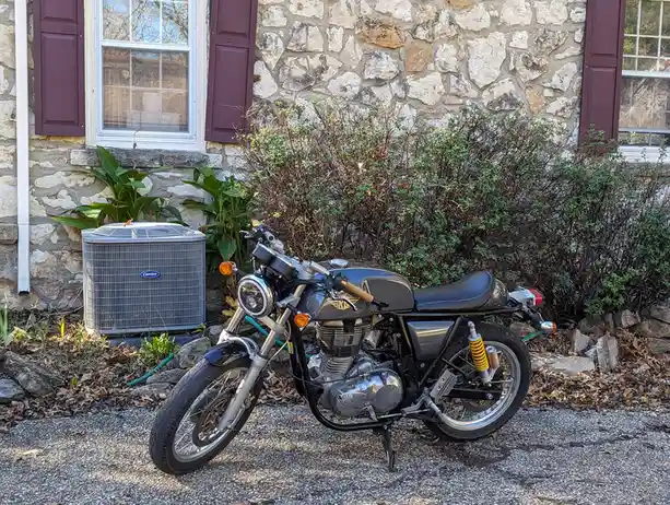 A black classic-style motorcycle with chrome accents and yellow rear suspension is parked on gravel in front of a stone…