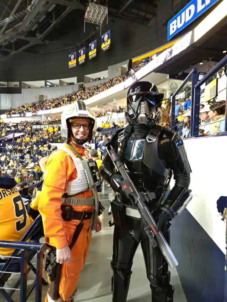 Two people in Star Wars costumes pose at a crowded sports arena; one is dressed as a Rebel pilot in orange flight gear and…
