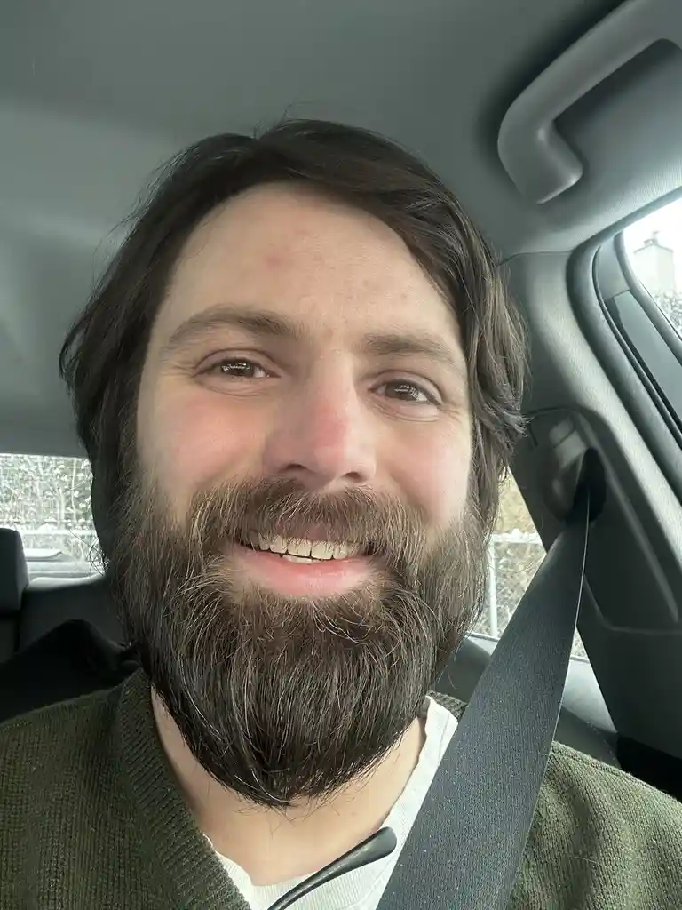 A man with a full beard and brown hair smiles at the camera while seated in a car, wearing a seatbelt over a green sweater…
