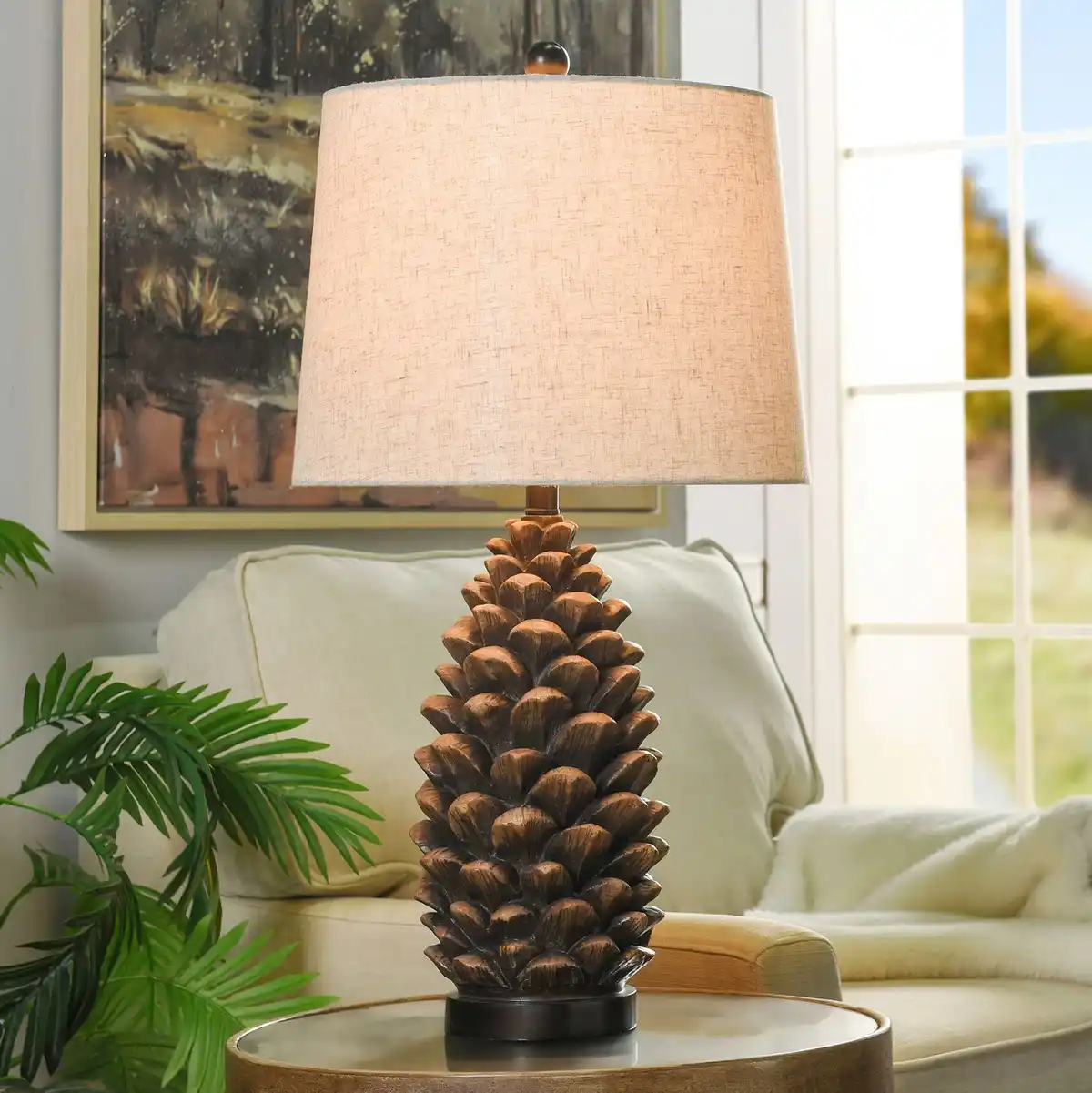 A lamp with a base shaped like a pine cone sits on a side table in a living room, with a beige lampshade, a couch, a plant…