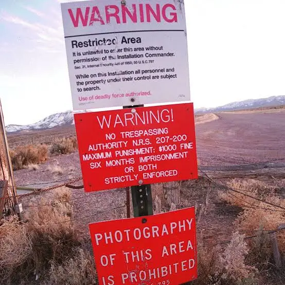 Three stacked warning signs in a barren, desert-like landscape with mountains in the background. The top sign says 'WARNING…