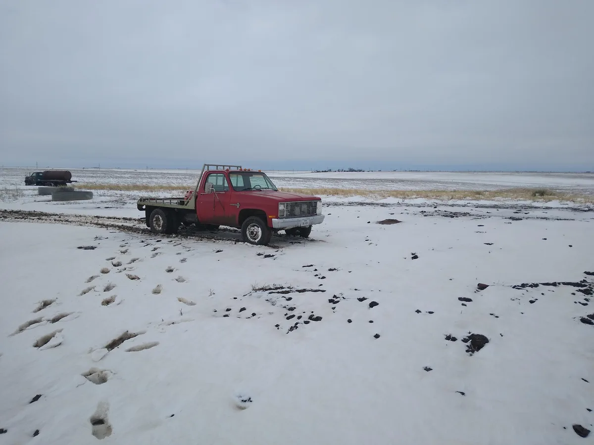 A red flatbed pickup truck is parked in a snowy, open field with visible tire tracks and footprints in the snow. In the…