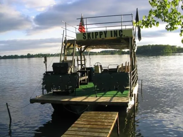 A rustic, green-painted flat-bottomed boat with a wooden dock attached, floating on calm water under a partly cloudy sky…