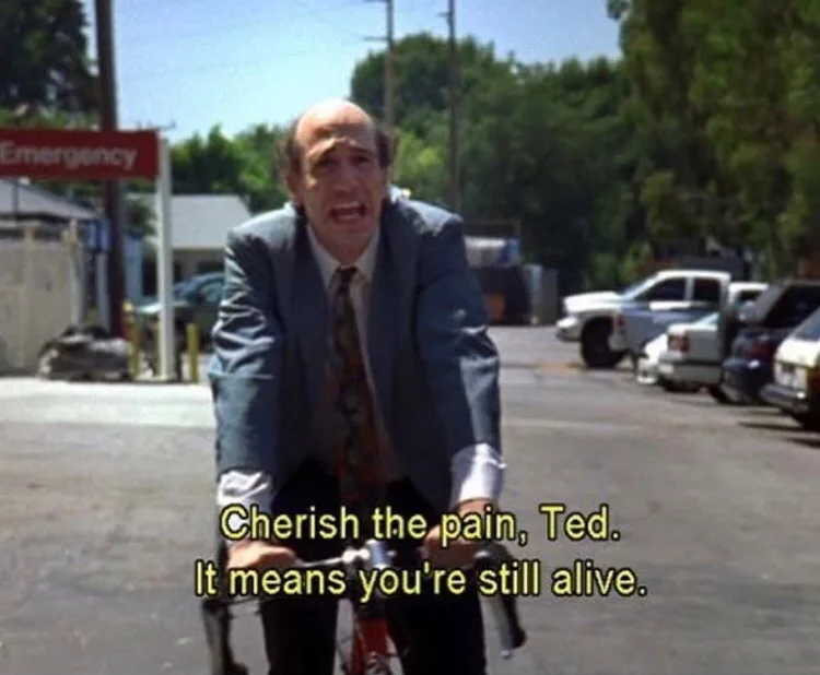 A man in a suit and tie is riding a bicycle outdoors with a strained, pained expression. Behind him, an 'Emergency' sign is…