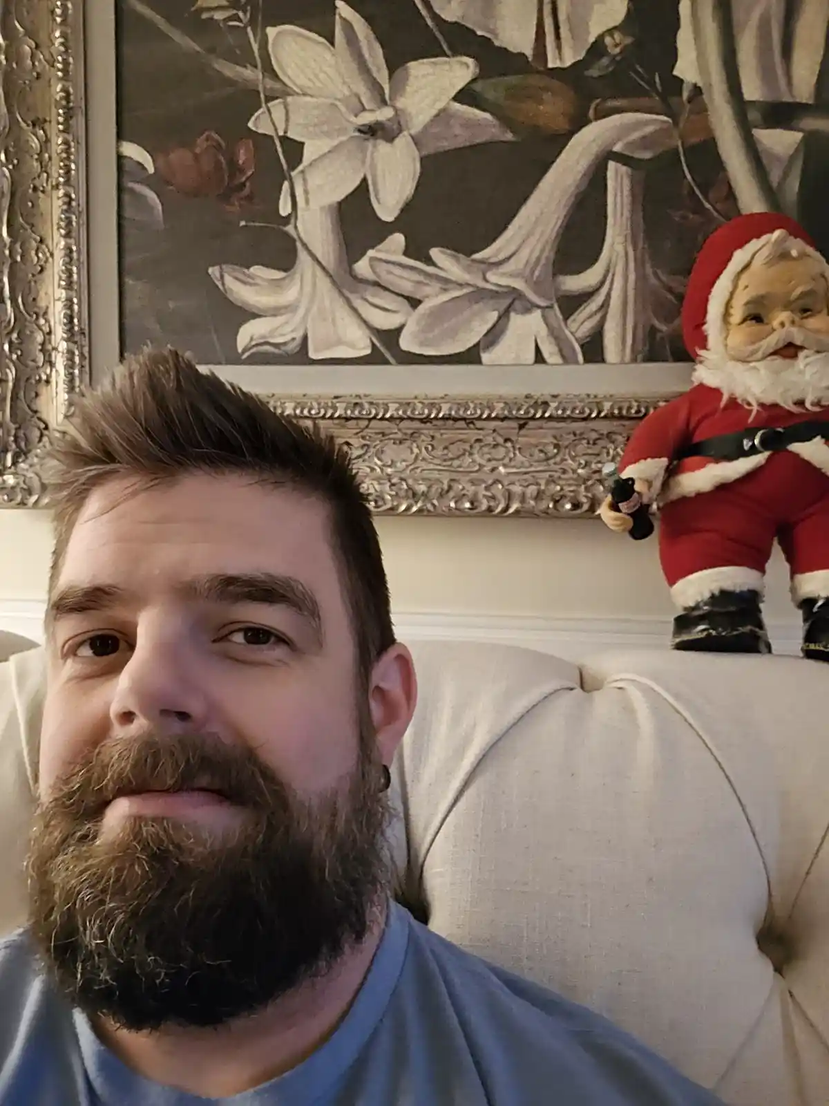 A man with a beard sits on a couch, looking at the camera, while a small Santa Claus figurine wearing a red suit and holding…