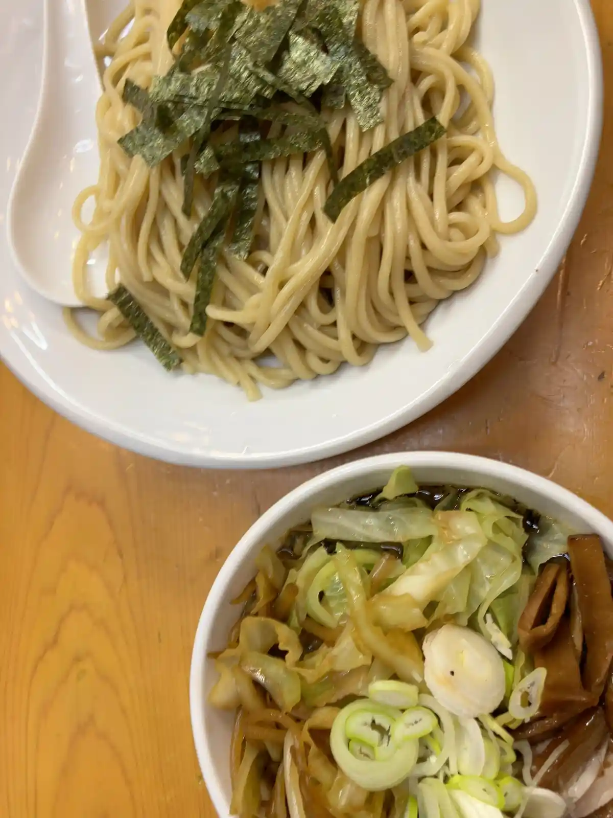 A close-up photo shows two Japanese noodle dishes on a wooden table: a white plate with soba noodles topped with green nori…