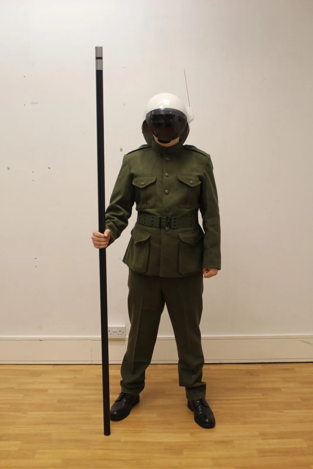 A person in a full green military-style uniform and a white riot helmet with a dark visor stands indoors holding a tall…