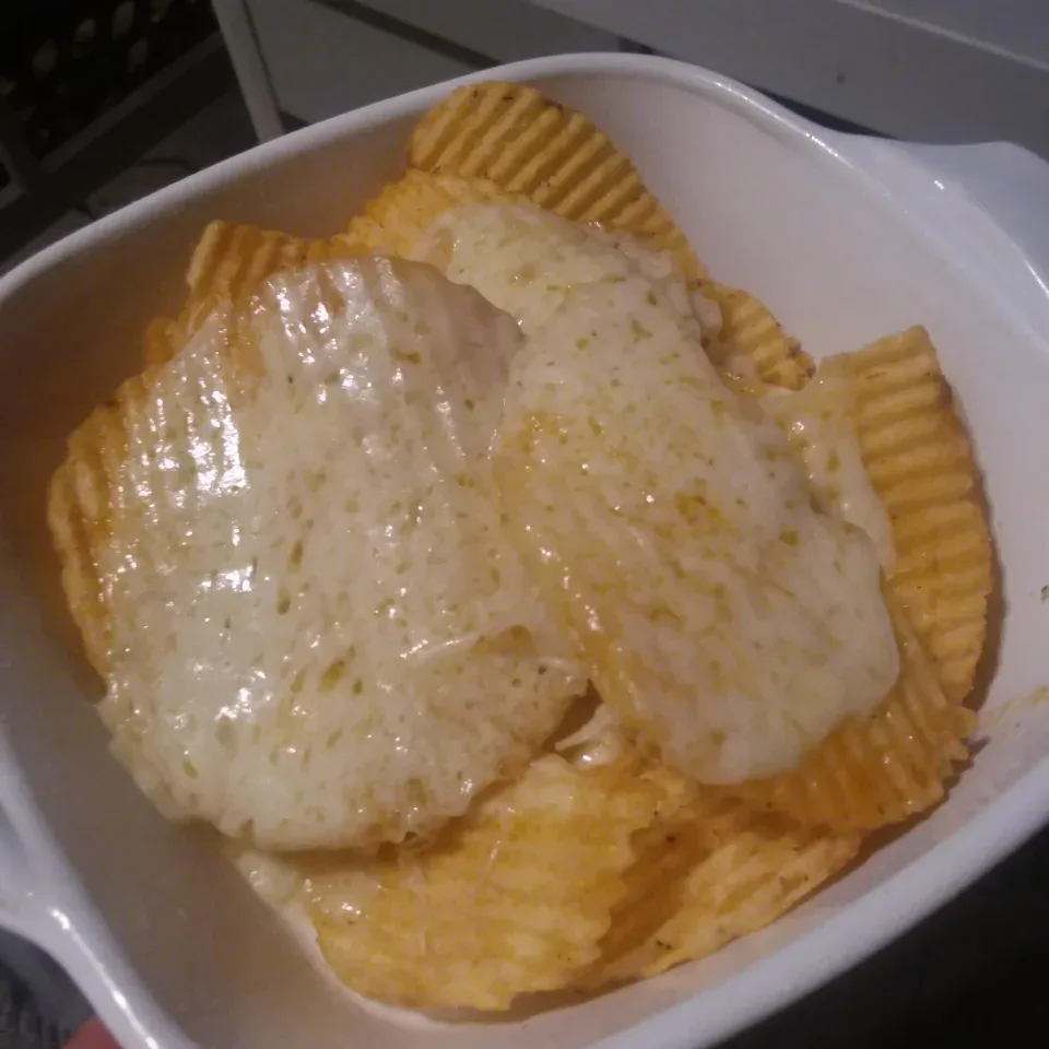 A white ceramic dish holds ridged, golden-brown potato chips generously topped with a thick, melted, slightly bubbly layer…