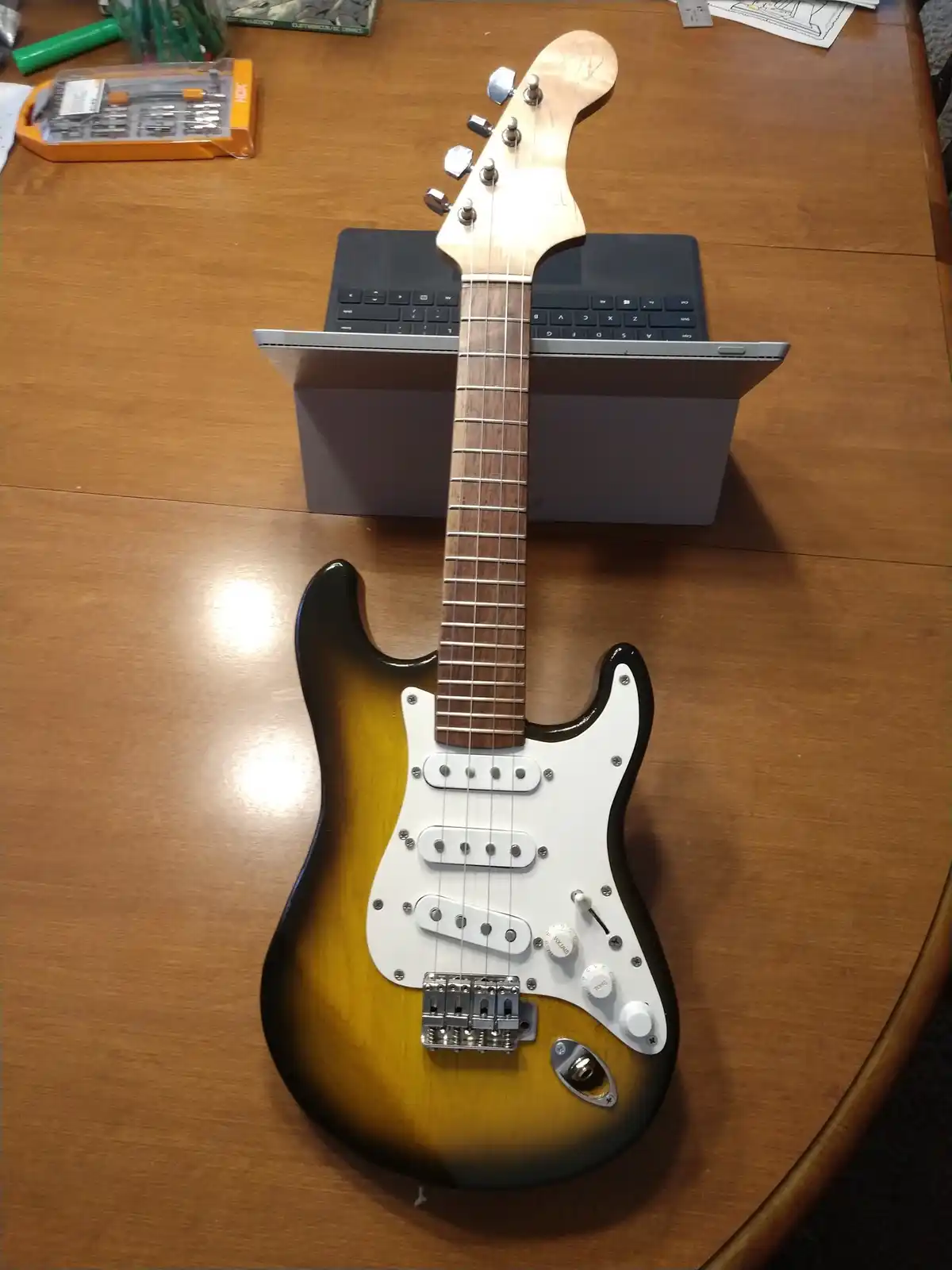 A sunburst-finish electric guitar with a white pickguard is resting on a wooden table, its neck propped up against the open…