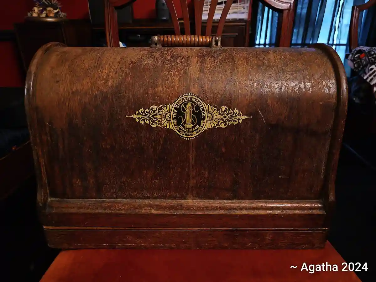 A close-up of a vintage, worn wooden suitcase with a brass 'The Singer Manfg Co Trade Mark' emblem, sitting on a red chair…