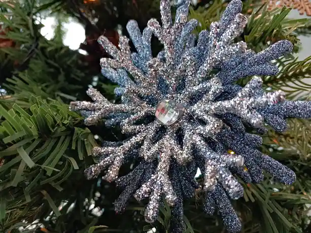 A close-up photo shows a glittery, blue-and-silver snowflake ornament hanging on a green artificial Christmas tree, with a…