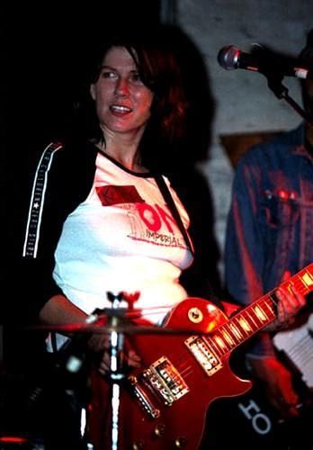 A woman with dark hair, wearing a white t-shirt with red and black graphics, plays a red electric guitar on stage under dim…
