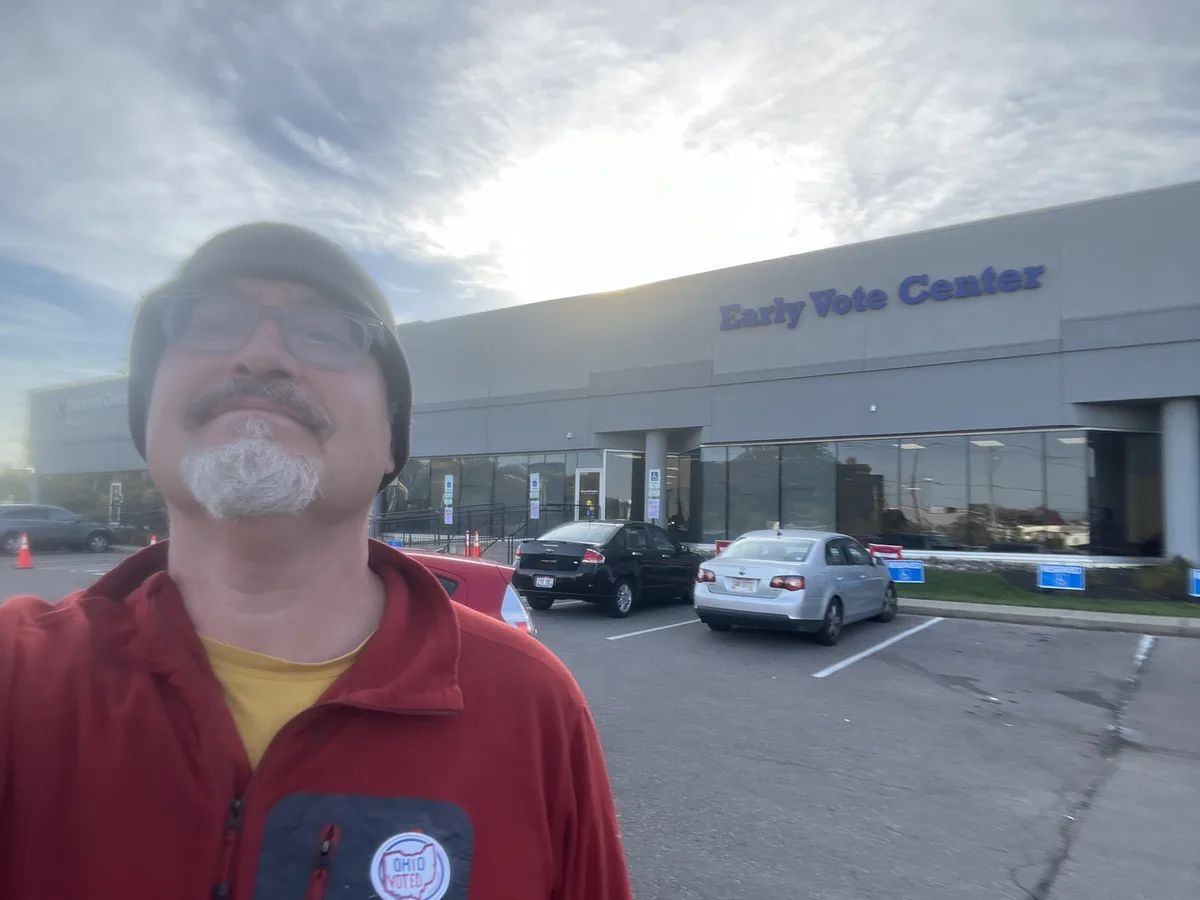 A man with a white goatee, glasses, and a beanie stands in front of an 'Early Vote Center' building, wearing a red jacket…