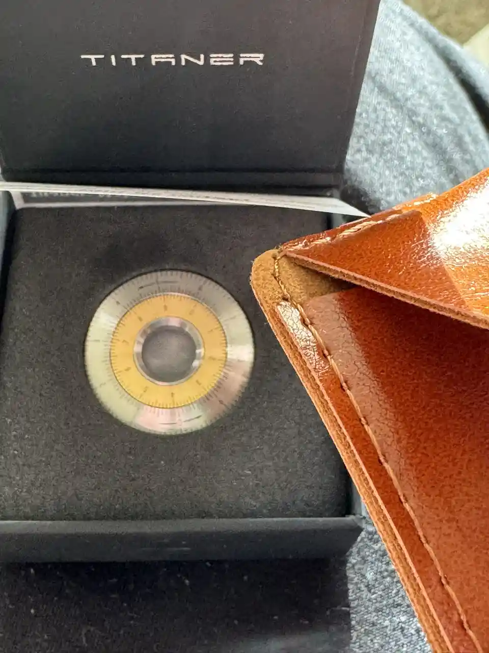 A close-up photo shows a circular, metallic watch dial with a yellow center and measurement markings, resting in a black…