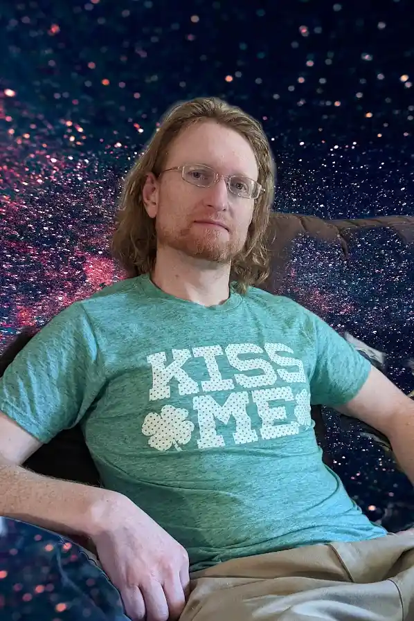 A man with glasses and a beard sits on a couch wearing a green t-shirt that says 'KISS ME' with a shamrock replacing the…