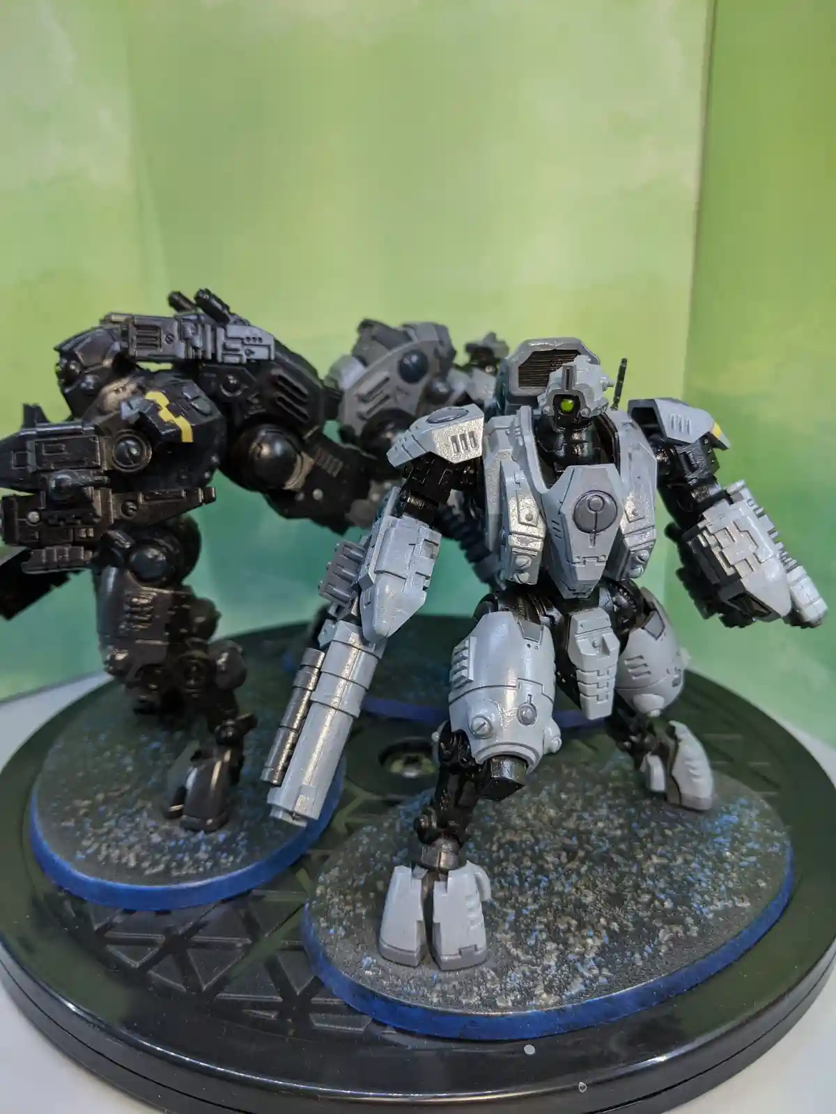 Two detailed, painted mecha figurines stand on circular bases against a green background. The foreground mecha is silver and…