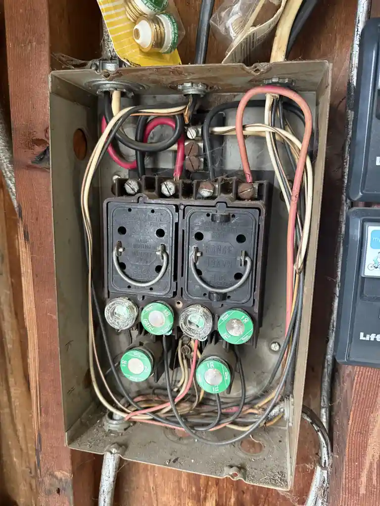 A close-up photo of a dusty, old, and messy electrical fuse box with tangled wires, spiderwebs, and green fuses. The box has…