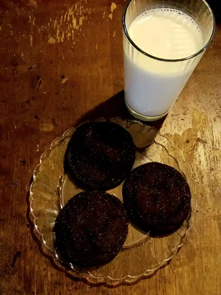 A top-down photo shows three dark chocolate cookies on a clear glass plate next to a tall glass of milk, both resting on a…