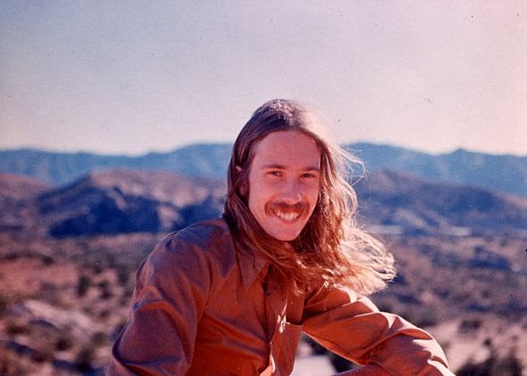 A vintage photo of a smiling man with long hair and a mustache, wearing a brown shirt, posing outdoors with mountains in the…