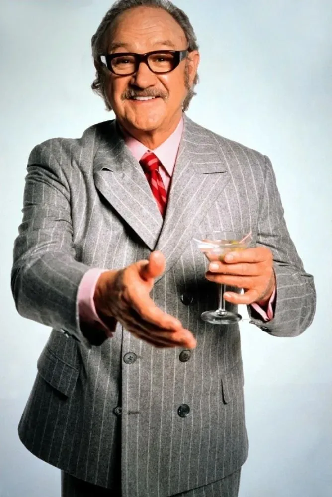 a man in a grey pinstripe suit, pink shirt, and red tie holds a martini glass in one hand while extending the other in a…