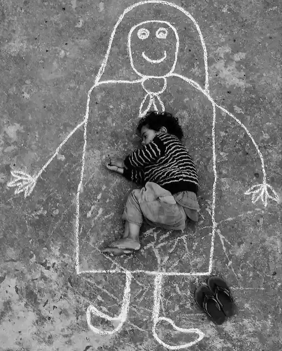 A black-and-white photo shows a young child sleeping inside a large chalk drawing of a smiling person on a concrete surface…