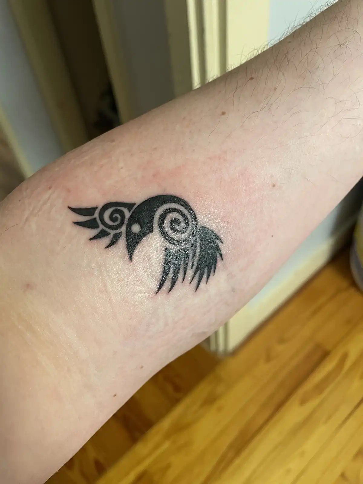 A close-up photo shows a black ink tattoo on a person's inner forearm, depicting a stylized bird with spiral patterns on its…