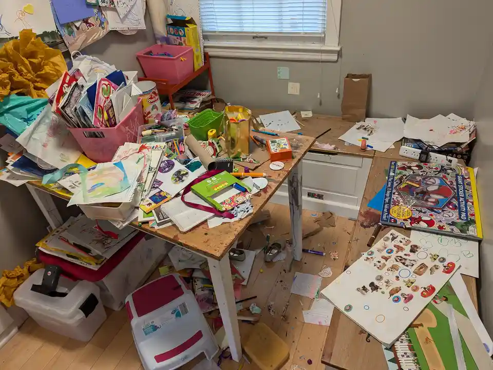 A messy craft room with a wooden table overflowing with art supplies, drawings, stickers, and scattered papers. The floor is…