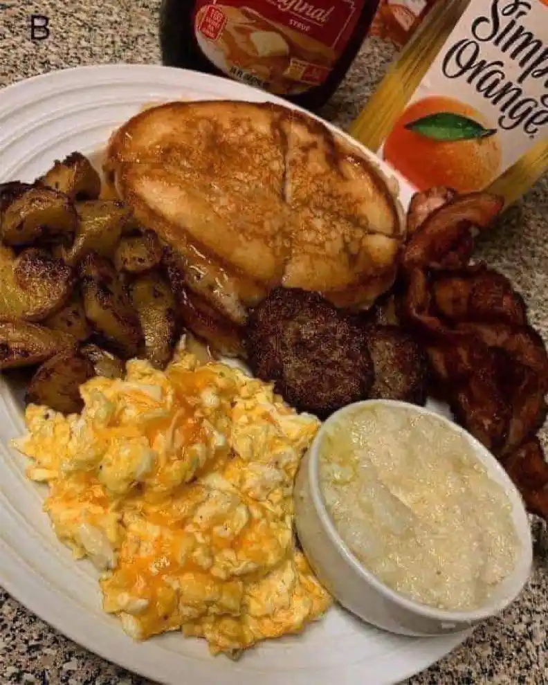 A plate of breakfast food including scrambled eggs with cheese, roasted potatoes, bacon, sausage patties, a grilled…