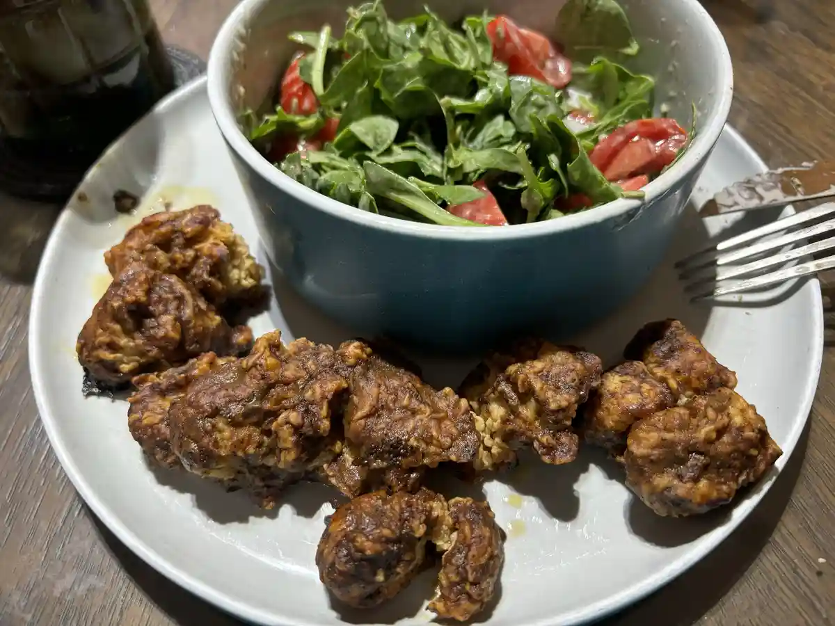 A white plate holds a blue bowl of salad with greens and tomato slices, alongside several pieces of fried, breaded, and…