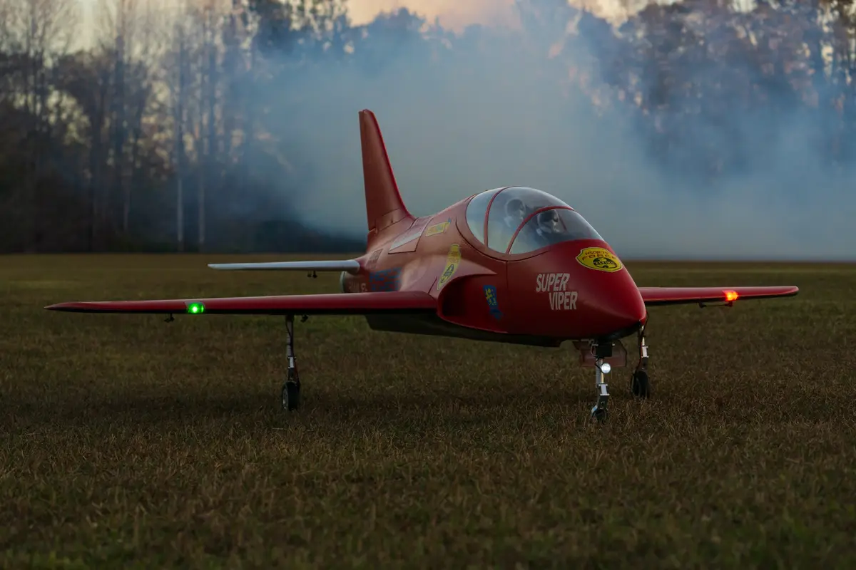 A red model jet airplane, labeled 'SUPER VIPER' on its nose, sits on a grassy field with smoke in the background. The…