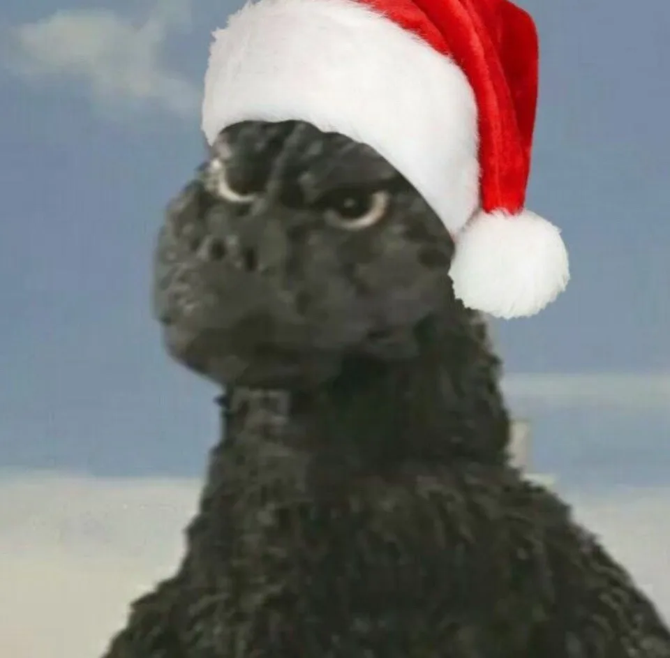 A close-up of the monster Godzilla wearing a red and white Santa hat, looking upward with a neutral expression against a…