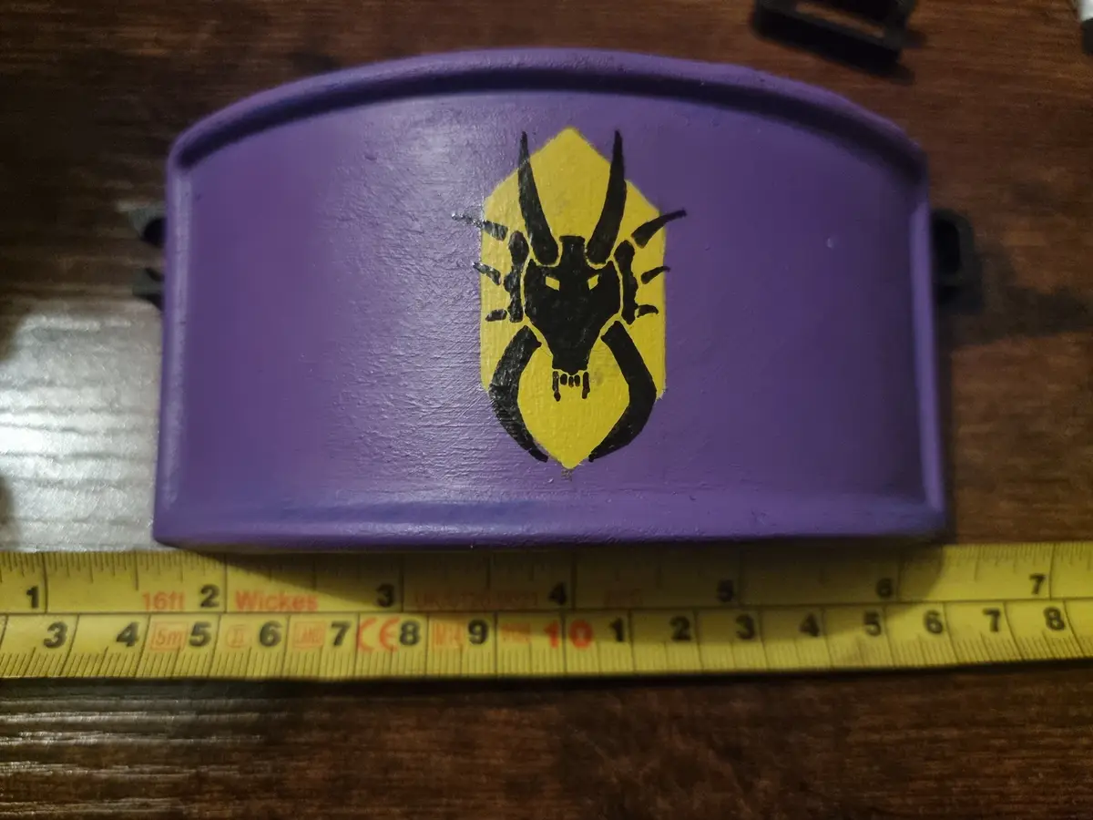 A purple plastic object, resembling a toy or small container, features a hand-painted black spider emblem on a yellow…