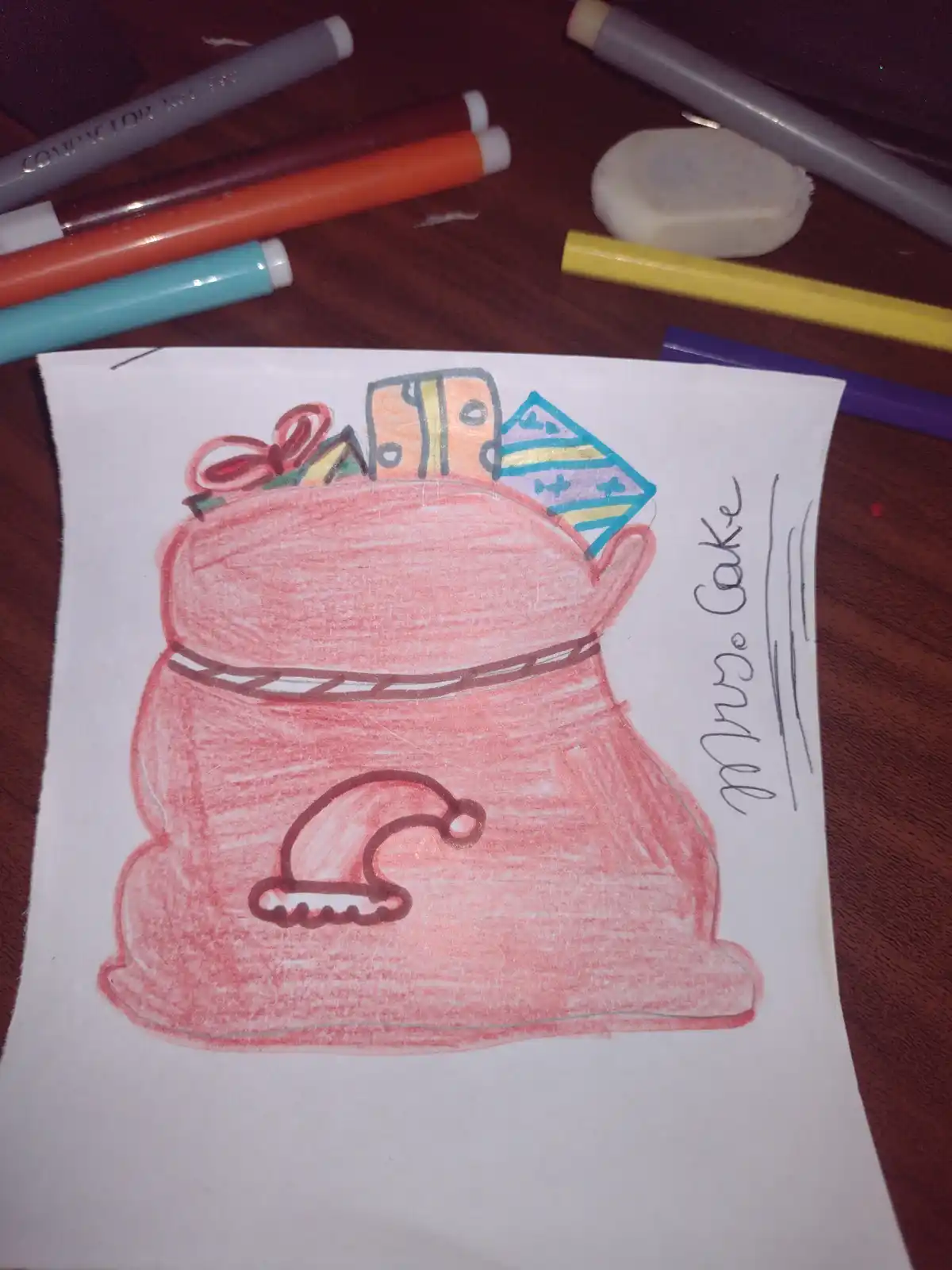 A child's crayon drawing shows a red sack overflowing with colorful presents and a Santa hat, with the handwritten text…