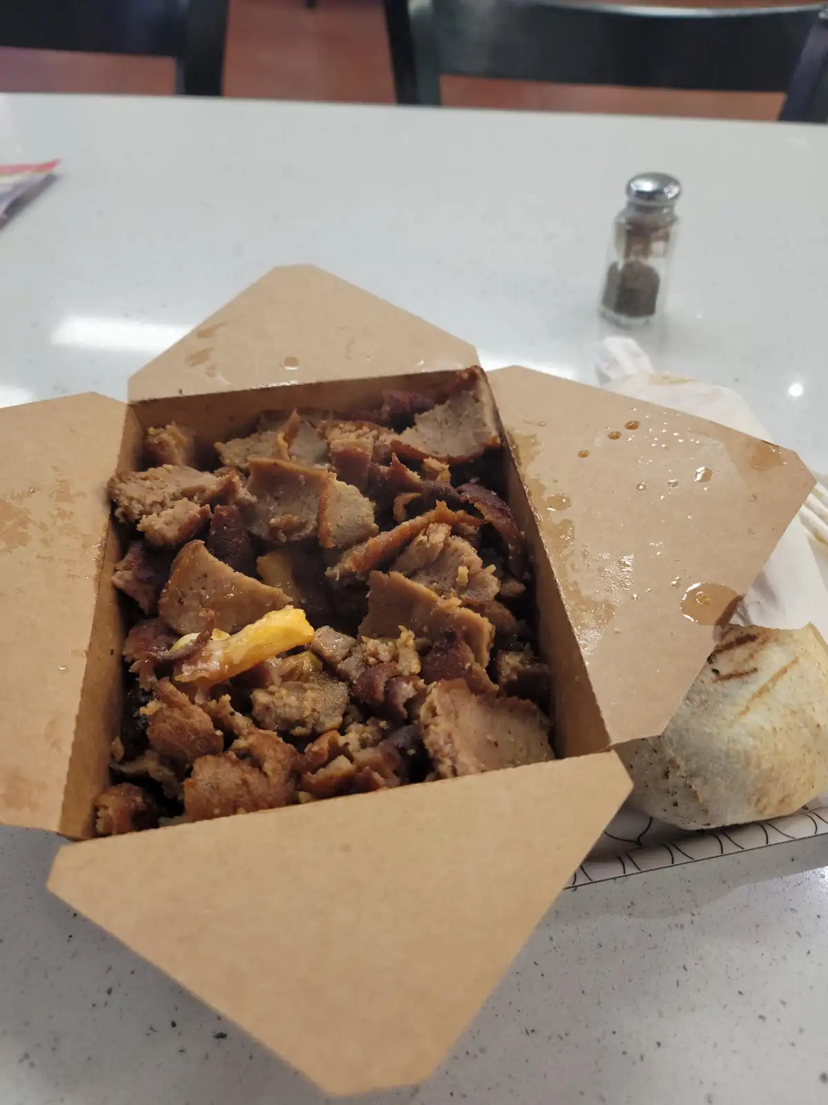 A cardboard takeout container filled with chopped, crispy meat and a single yellow cheese slice sits on a white speckled…