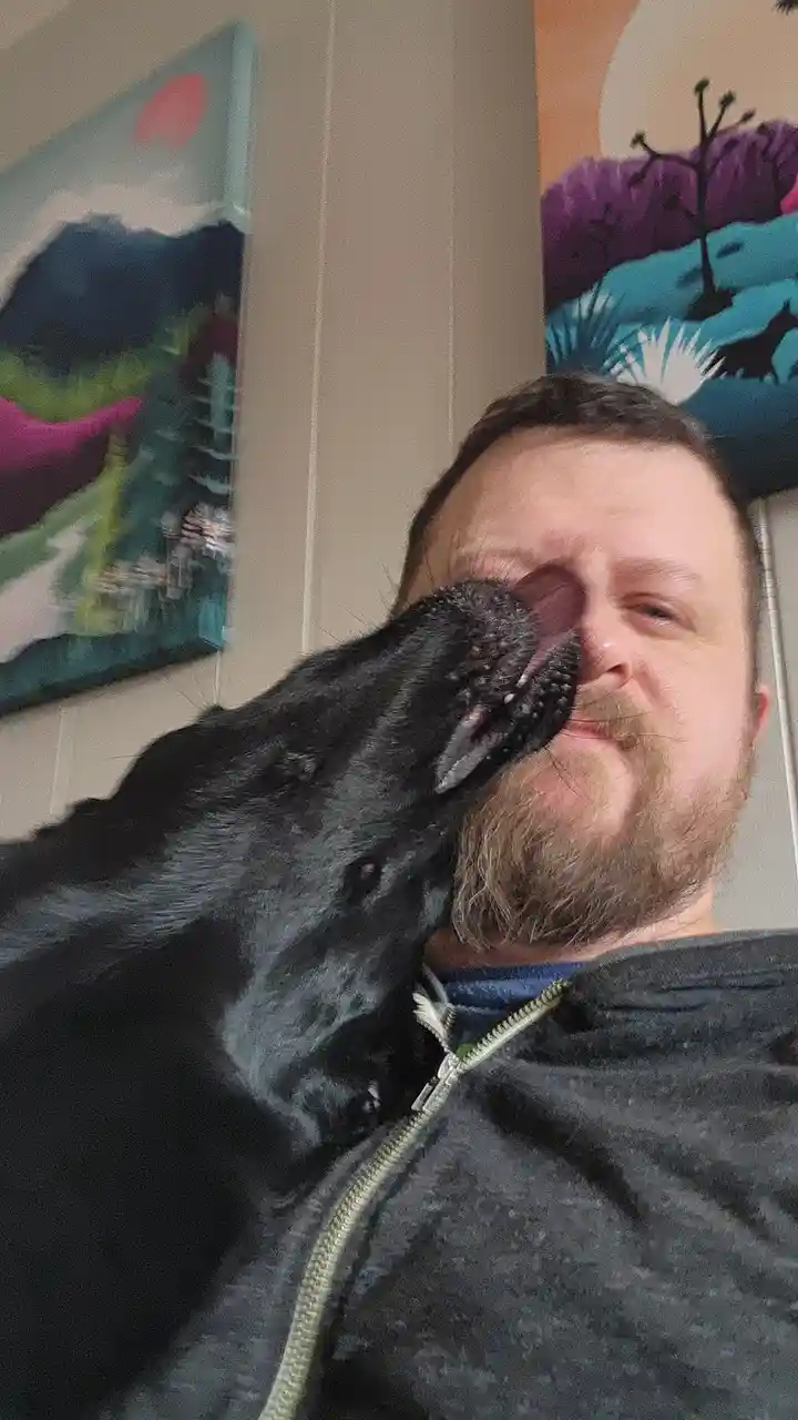 A man with a beard and a dark zip-up hoodie is taking a selfie, and a black dog is licking his face. The dog's snout is…