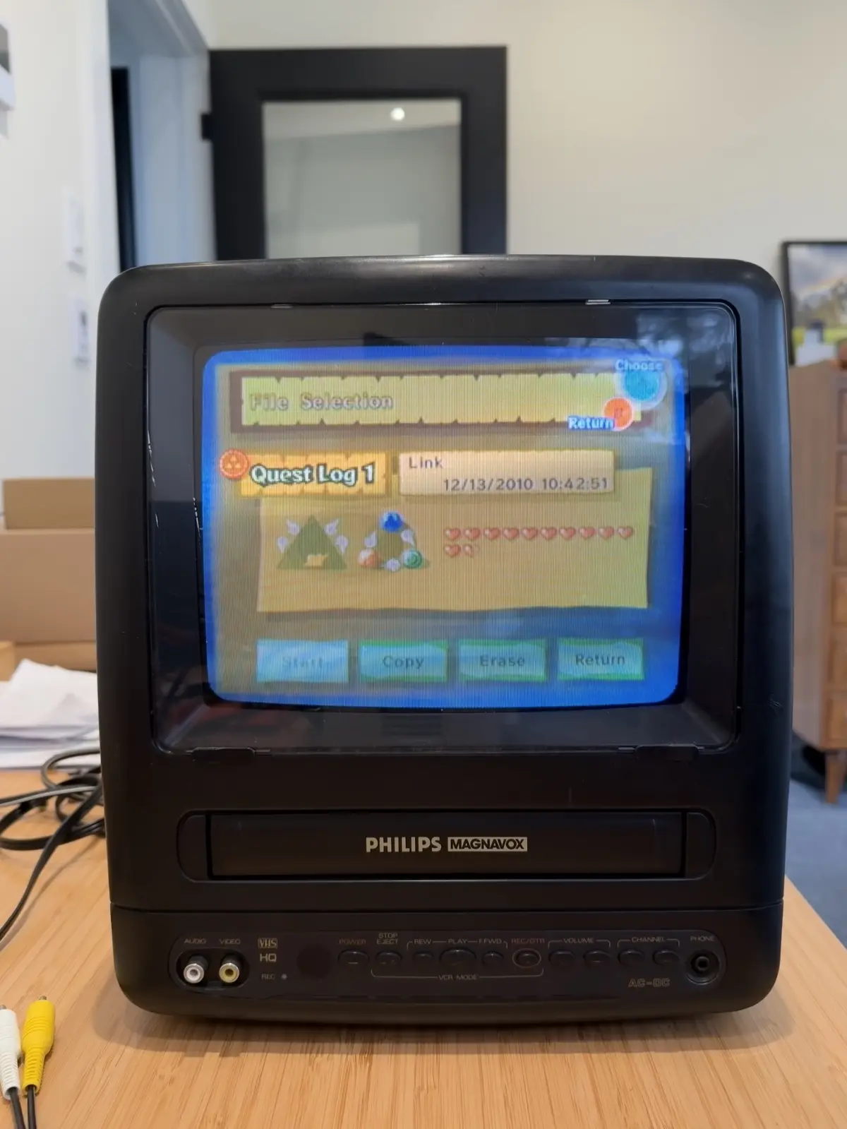 A black Philips Magnavox VCR/TV combo unit displays a video game menu titled 'File Selection' with 'Quest Log 1' selected…