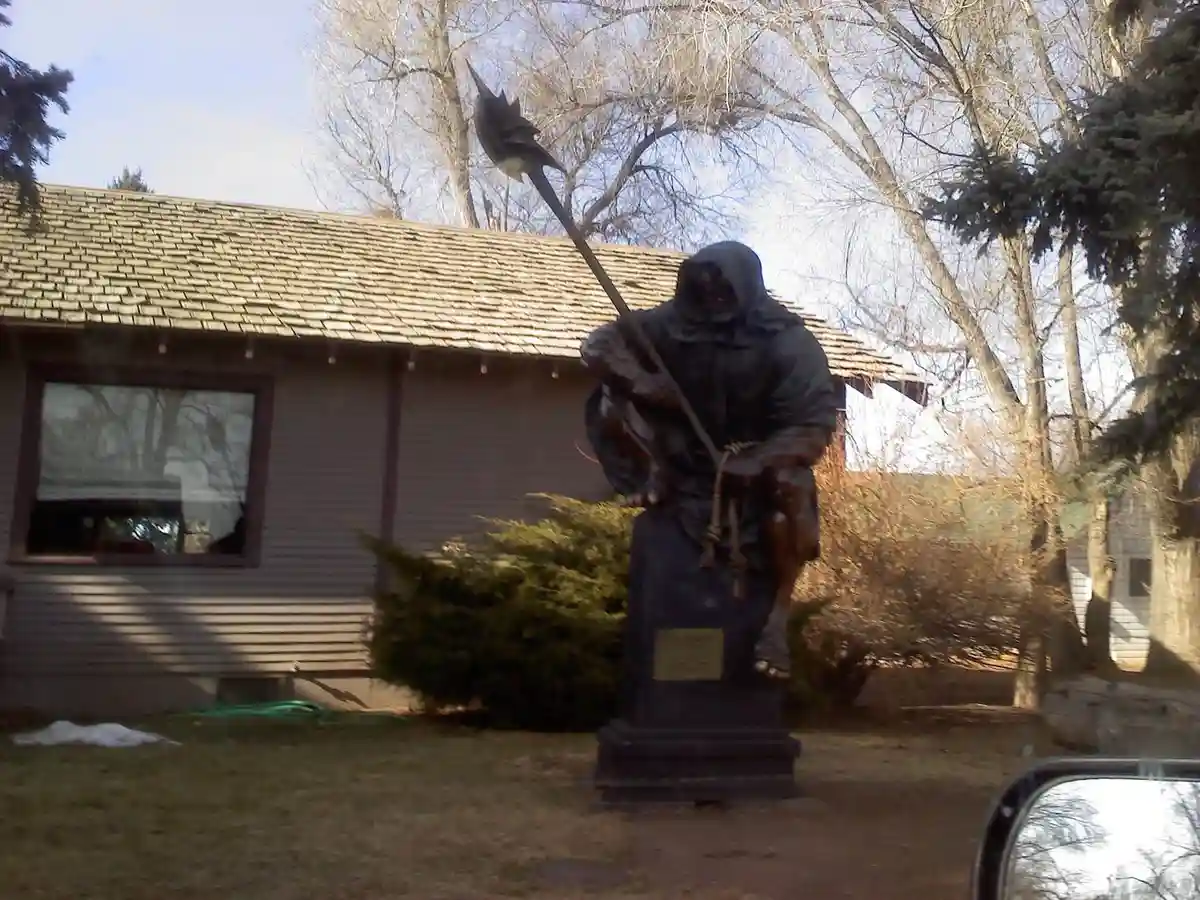 a bronze statue of the grim reaper, hooded and holding a scythe, is seated on a stone bench in a residential yard. Behind it…