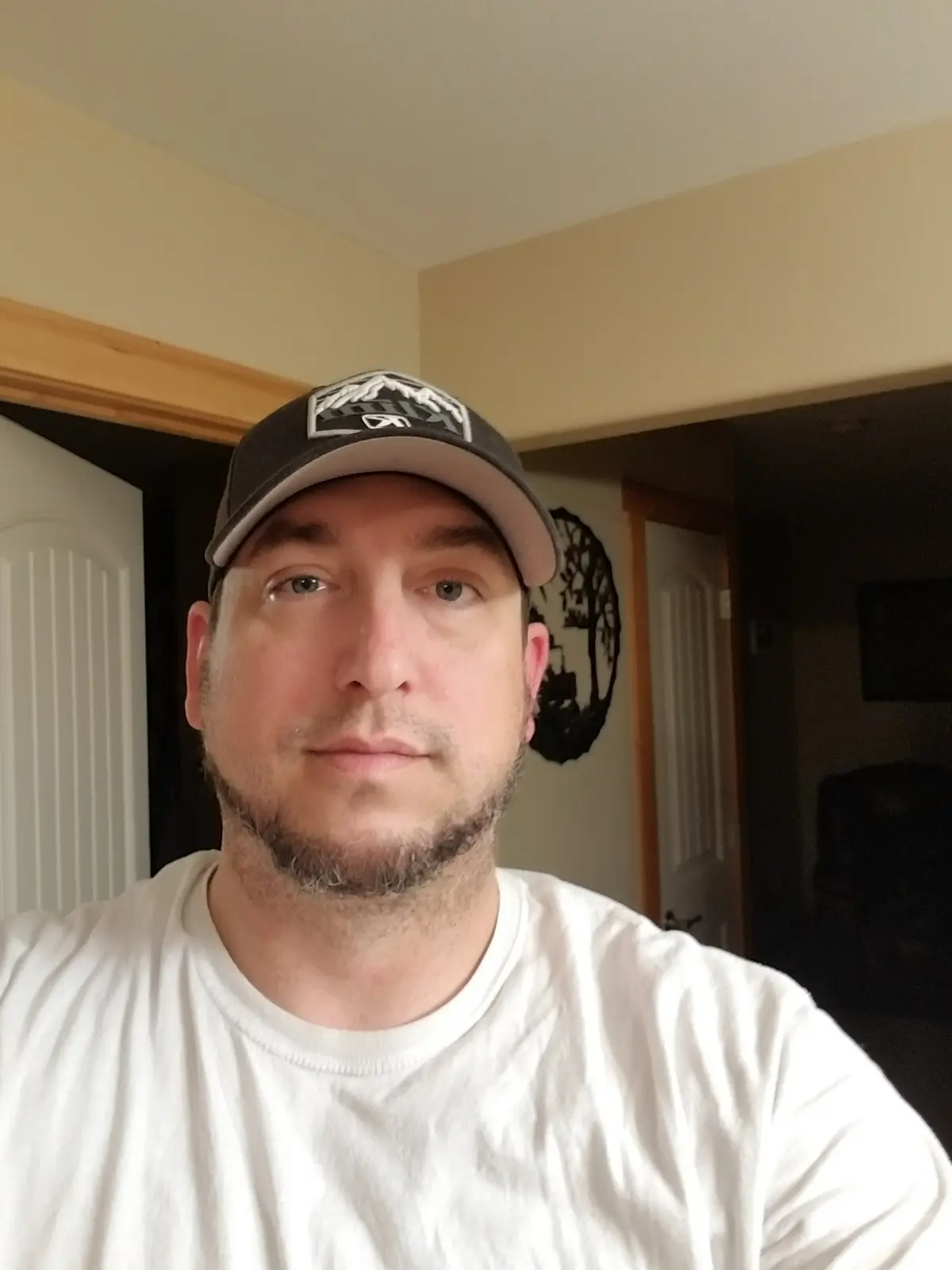 A man with a beard and a baseball cap looks directly at the camera with a neutral expression, wearing a white t-shirt in a…